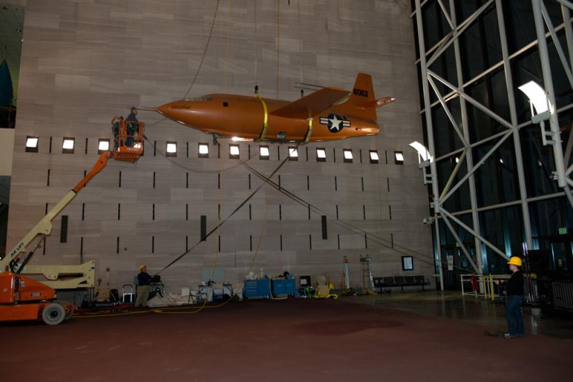 Museum employees in the middle of lowering the Bell X-1, an orange military monoplane, from the ceiling to the floor of the Boeing Milestones of Flight Hall. The aircraft is partially lowered.