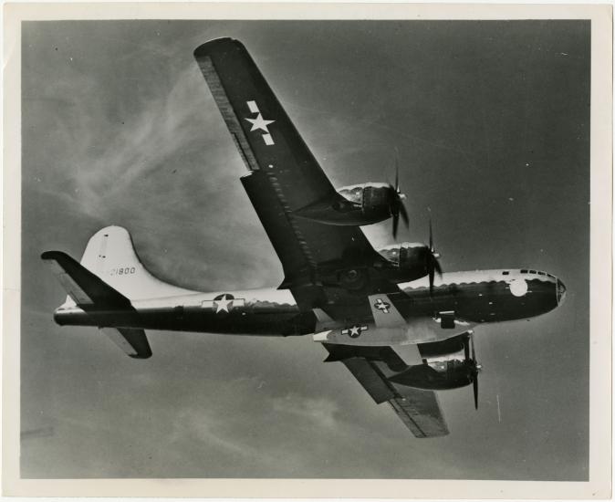 Bottom view of the Bell X-1, a military aircraft, being lifted under a B-29, a larger military aircraft. The B-29 is in flight.