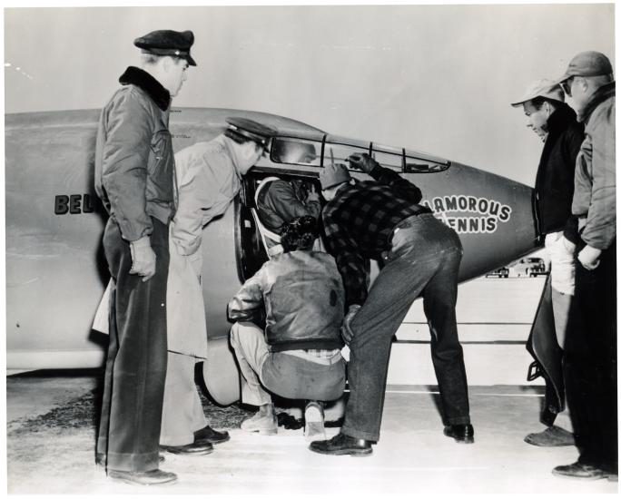 Chuck Yeager, a pilot, prepares for a flight in the cockpit of the Bell X-1 monoplane. Multiple people assist him prior to takeoff.