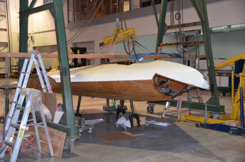 Diagonal side view of part of a white and wooden-colored glider aircraft, which is placed in between two metal frames. A section of clear plastic is removed from the front of the aircraft.