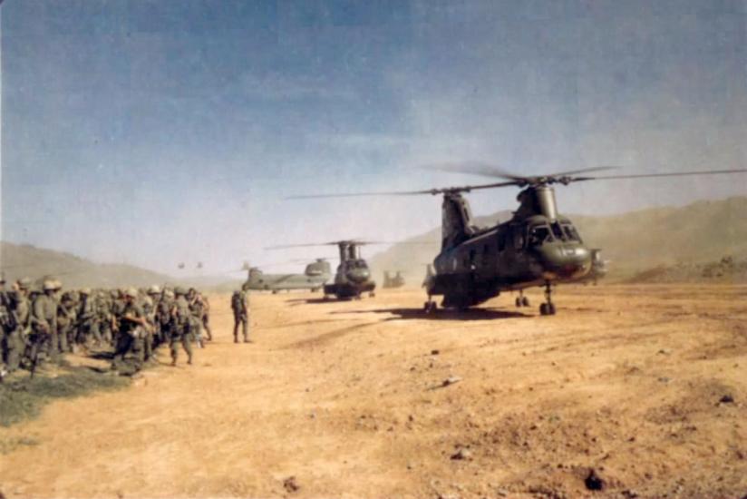 A group of soldiers stand near a sandy plain during the Vietnam War. On the sand, multiple military helicopters are visible and are stationary.