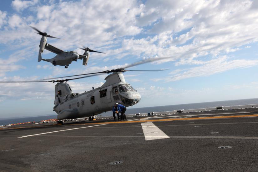 A helicopter with two sets of propellors placed on the sides of the aircraft flies over a helicopter on a tarmac with propellors on top of the body.