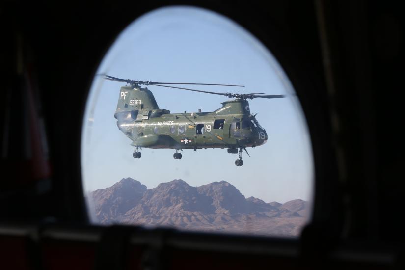 Porthole-style view of green military helicopter with two sets of propellors on top of the helicopter's body. The helicopter is in flight.