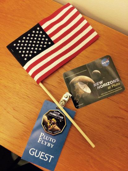 An American flag, blue guest badge, and sticker featuring Pluto that were given out at an event celebrating the New Horizons spacecraft.