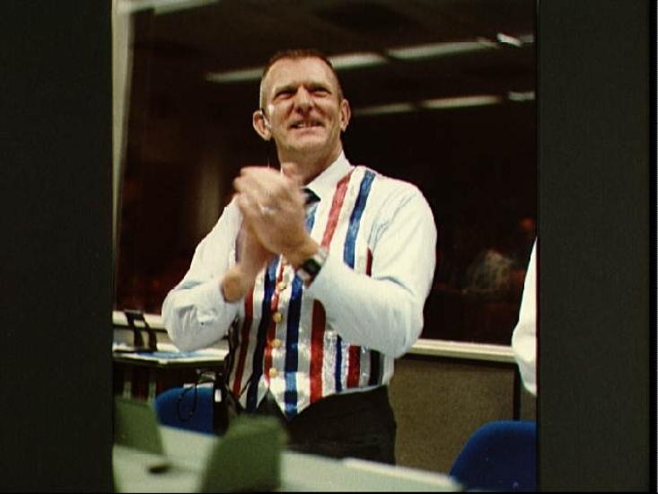 Eugene C. Kranz, a white male who served as Director of Missions Operations during Space Shuttle Mission STS-41C, wears a blue, red, and white vest in celebration. He is clapping.