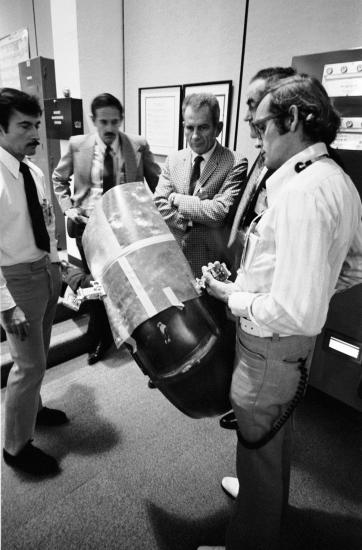 A group of five male NASA employees on Earth work together to come up with a solution for a wheel fender repair on the Lunar Module during the Apollo 17 mission.