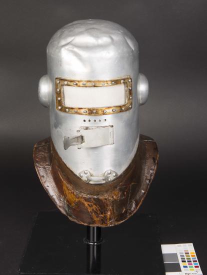 Frontal view of the first pressure helmet. The helmet is made from metal and is experiencing white-colored corrosion, requiring conservation treatment.