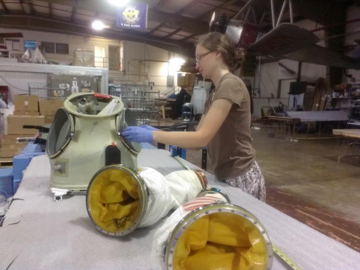 A white, female Museum intern works with spare spacesuit parts including arm sleeves and the neck piece.