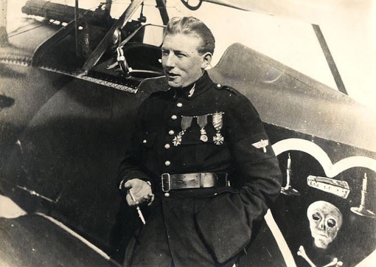 Charles Nungesser, a French World War I male pilot, poses in front of his aircraft and next to his famous insignia on the aircraft. The insignia consists of a skull, crossbones, coffin, and two candles inside a heart.