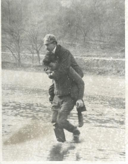 A mechanic working with Orville Wright gives Orville a piggyback as they walk in a river.