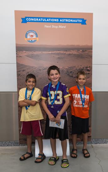 Three boys stand in front of a banner of Mars that is congratulating them on their completion of an astronaut game.