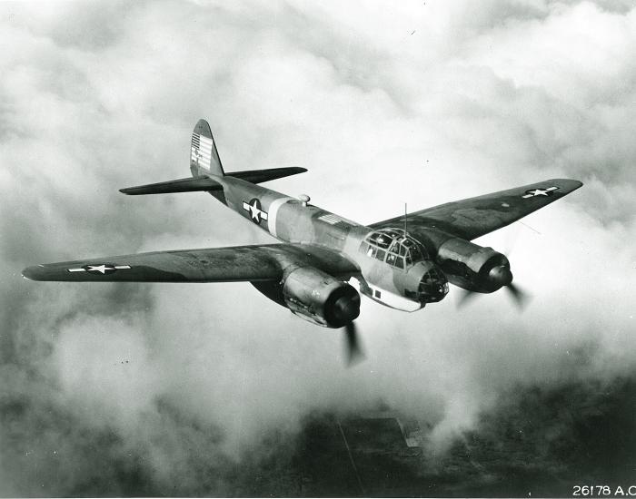 Diagonal front view of military monoplane with twin engines. U.S. star insignia is seen on the wings and rear fuselage. Aircraft is in flight.