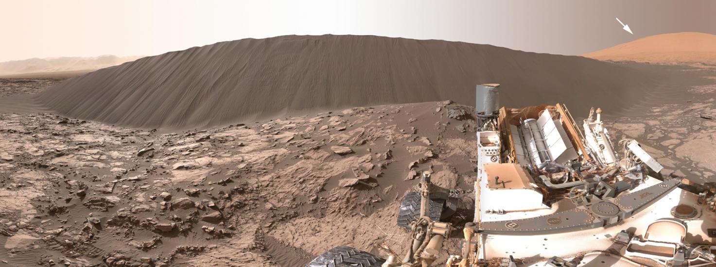 Tall sand dune in the background of a rocky, Mars surface. Curiosity Rover, a robotic surveyor, can be seen on the right side.