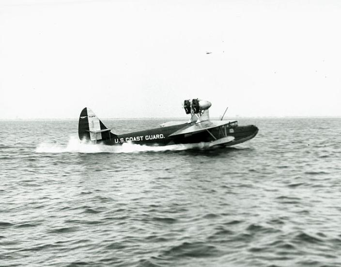 Side view of dark-colored sailplane with the name "U.S. Coast Guard" painted in a lighter color on the back fuselage of the aircraft. Two propellors can be seen on top of the front of the fuselage.