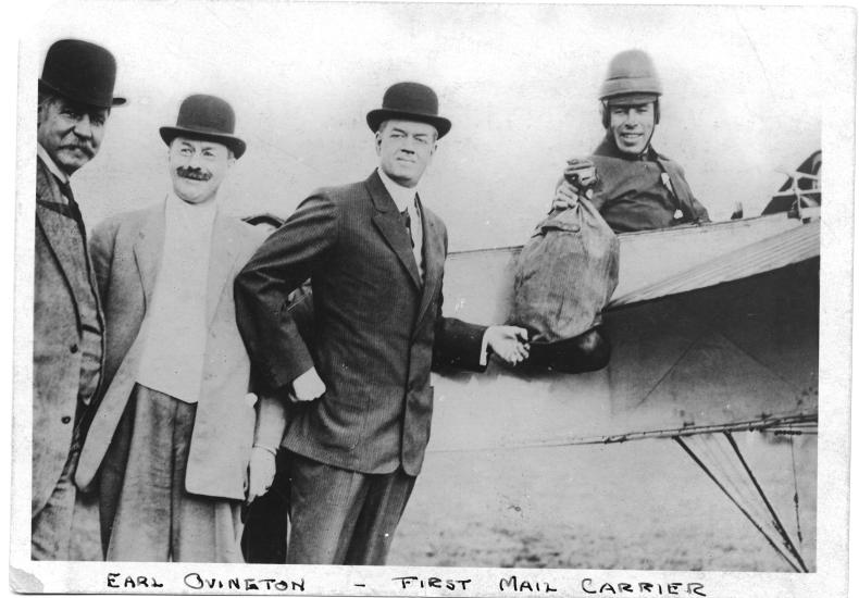 Earle Ovington, a male mail pilot, sits in the cockpit of an aircraft holding a bag of mail.