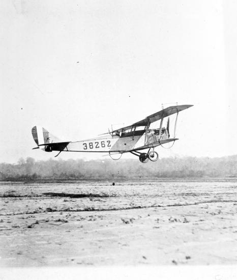 Side view of light-colored biplane in flight. Registration number "38262" written on back half of fuselage.