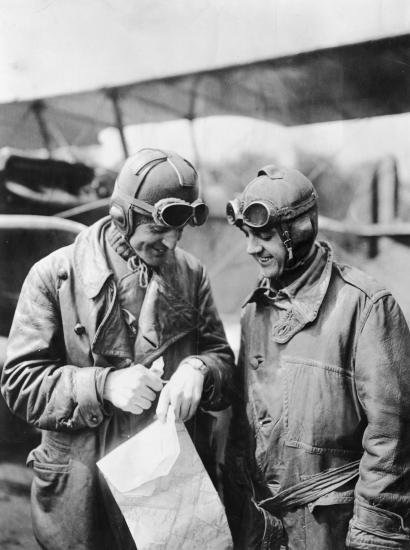 Two male military pilots in aviation gear look at a watch on the hand of the man on the left.