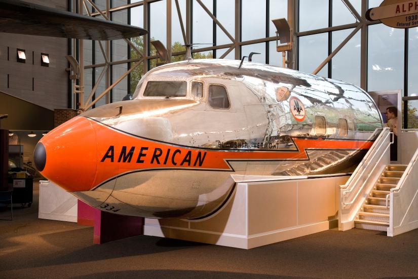 Nose, cockpit, and front fuselage section of a metal commercial airliner with orange, black, and silver-colored American Airlines livery.