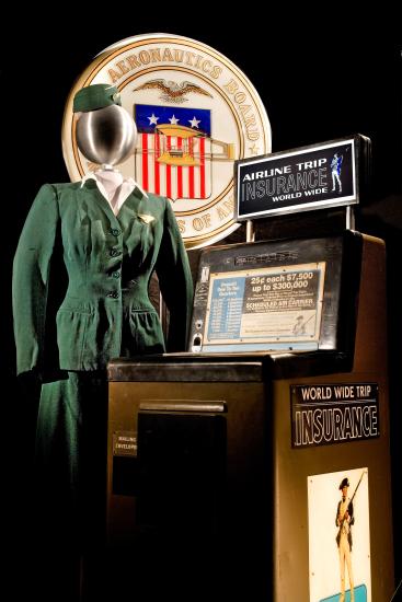 Brown-colored vending machine selling airline flight insurance. Next to the vending machine is a green flight attendant uniform styled on mannequin.