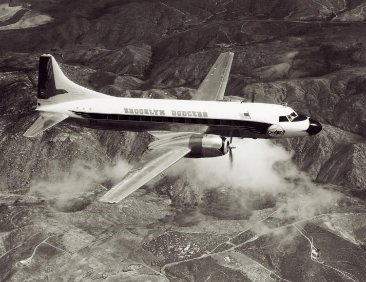 Side view of commercial aircraft in flight. Aircraft has two engines and features "Brooklyn Dodgers" name on the center of the side of the fuselage.