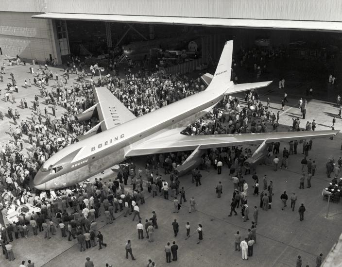 Top view of commercial jet with four engines and Boeing livery. People are crowded around the prototype jet.