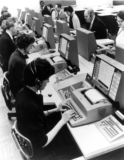 A group of people in an office setting use computers for airline reservations.