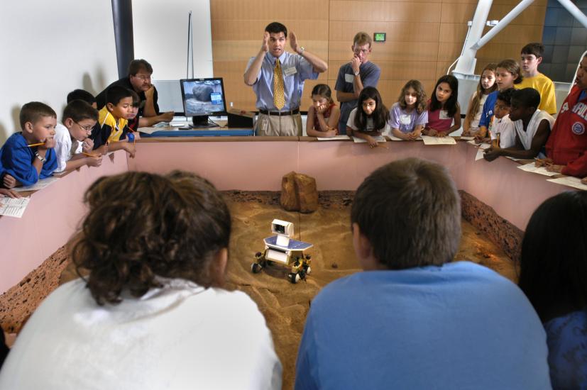 Museum visitors control a Mars rover model on a surface similar to the surface of Mars.