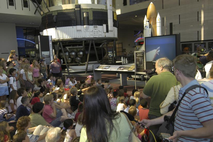 Museum visitors gathered around a large TV with Space Shuttle launch visible on the screen.