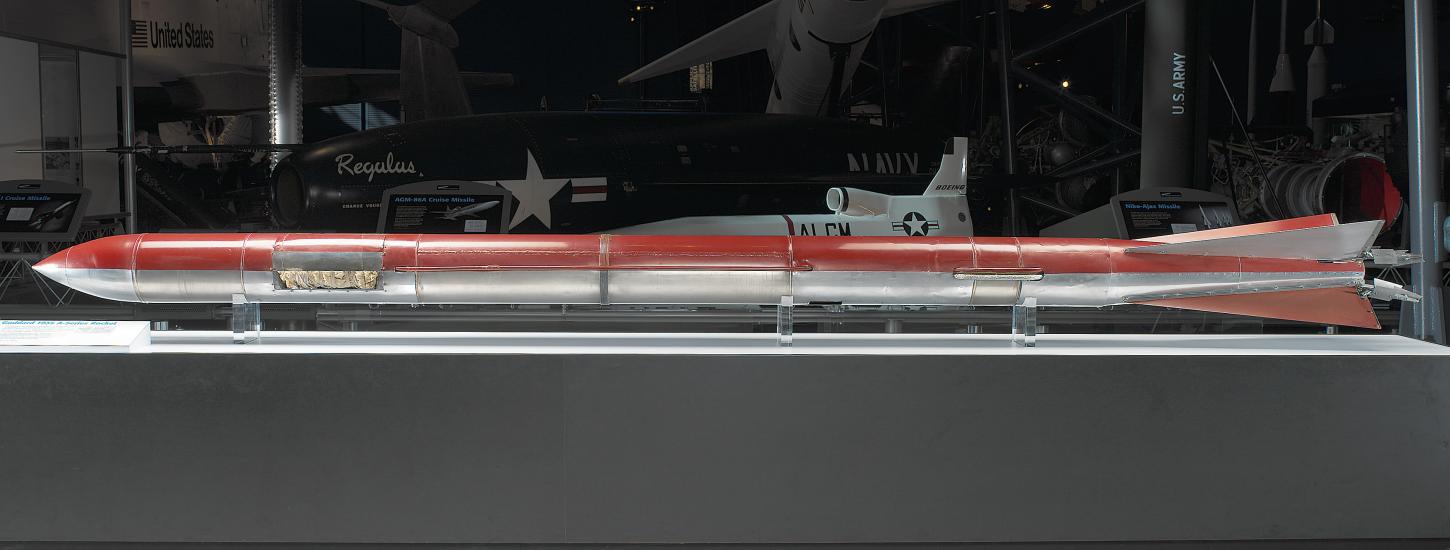 A long and skinny cylinder with a pointed tip at one end and tail fins at the other end. One half is painted red and the other half is left unpainted, showing the silver metal material it is constructed of.