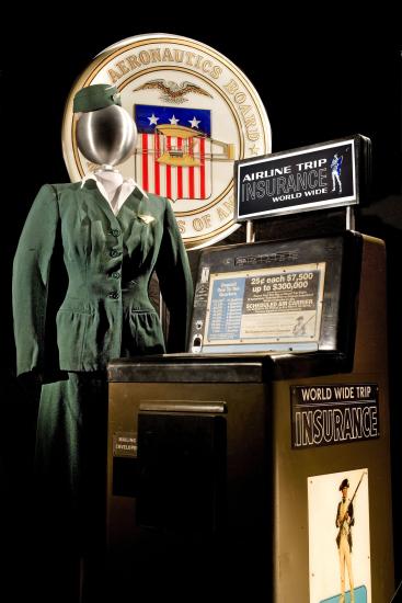 A mannoquin wears a 1950s green flight attendant's uniform next to an airline trip insurance machine.