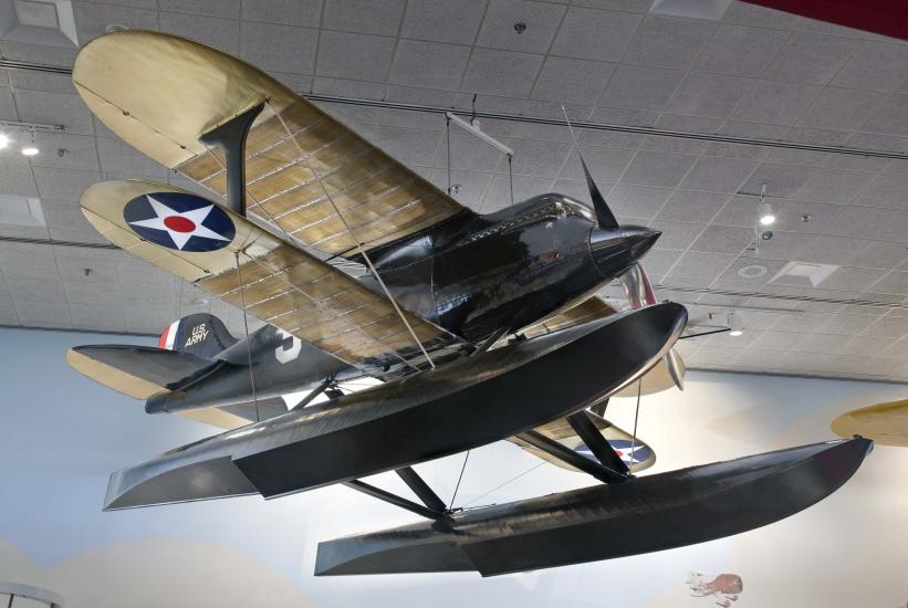 Black seaplane with pontoon floats instead of wheels and cream colored wings, hanging in museum