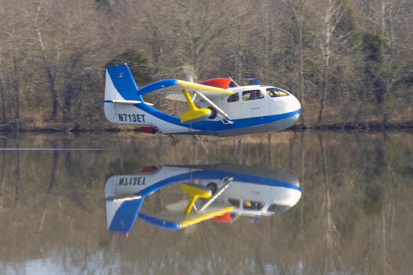 Side view of blue and white amphibious aircraft flying slightly above body of water. Registration number N713ET is painted in blue on the lower part of the vertical stabilizer.