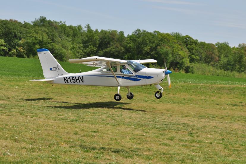 A white monoplane with a single engine flies just above the ground. The registration number "N15HV" is written in black near the rear of the fuselage.