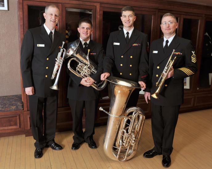 A set of four members of the U.S. Navy stand together in uniform, holding their respective brass instruments.