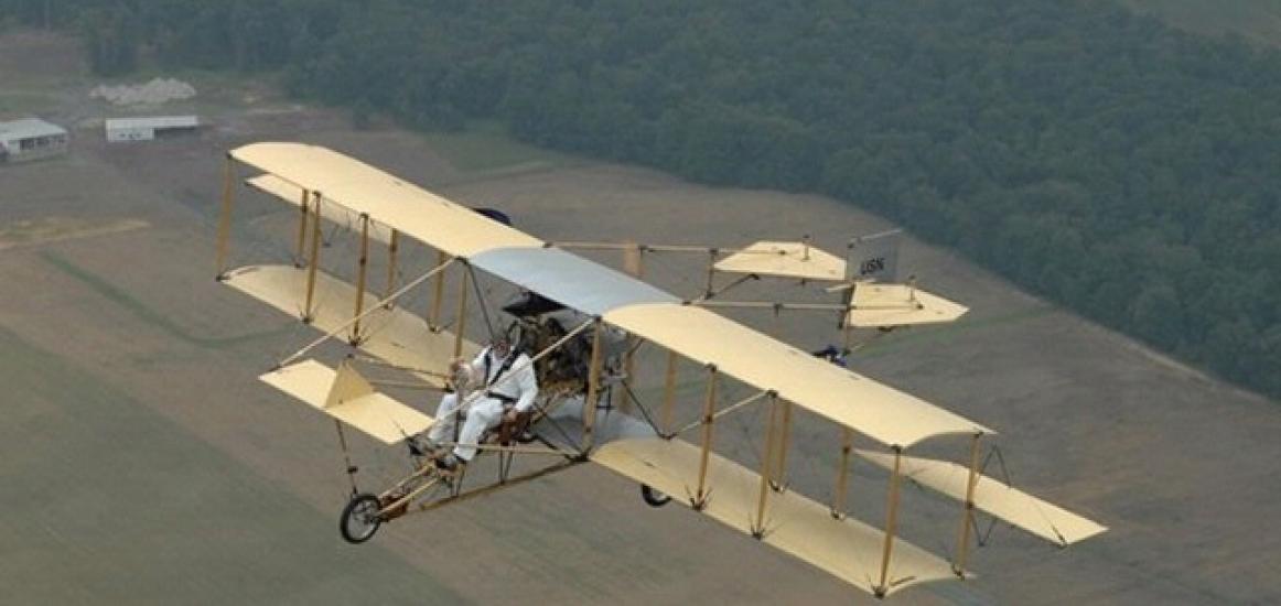 Diagonal side view of a biplane with yellow wings and a wooden frame during flight.