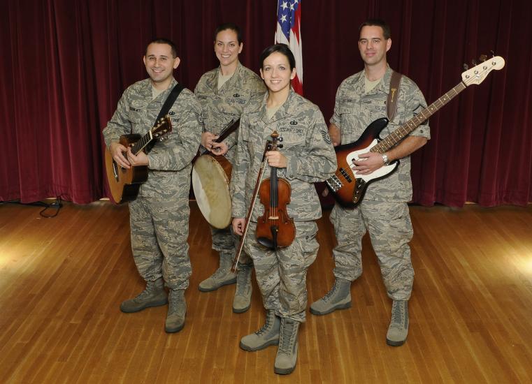 A group of four people pose together in U.S. Air Force camouflage uniforms. Each is holding an instrument: Two hold guitars, one plays a banjo, and the fourth plays the violin.