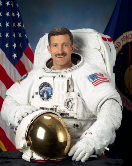 Dan Burbank, a male astronaut, stands for a photo wearing astronaut gear. He is standing in front of a background featuring a U.S. red, white, and blue flag and a second flag featuring NASA's seal.