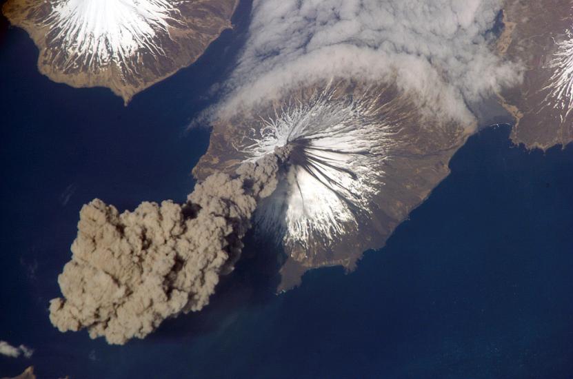 View from space of a volcanic eruption. A gray cloud of ash seen in the bottom left corner is floating away from the volcano site.