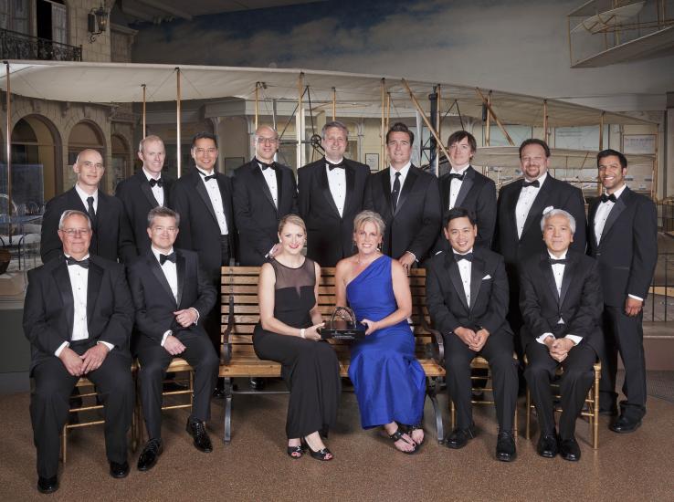 A large group of people sit and stand for a group picture after receiving a trophy from the museum. Two members sitting in the center hold the trophy.