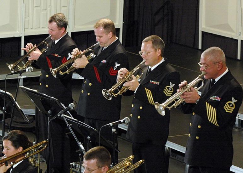 A group of musicians in U.S. Navy uniforms perform using various brass instruments.