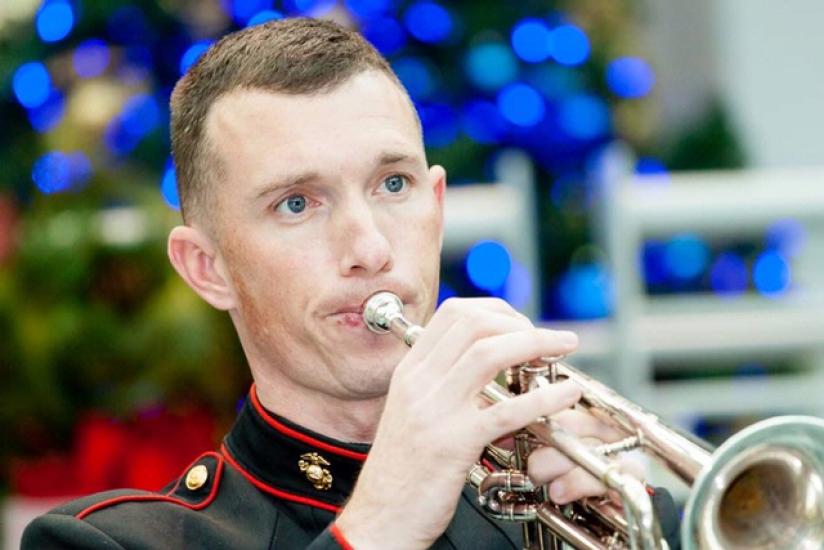 A man plays a trumpet in Marine Corps uniform as part of a larger group.