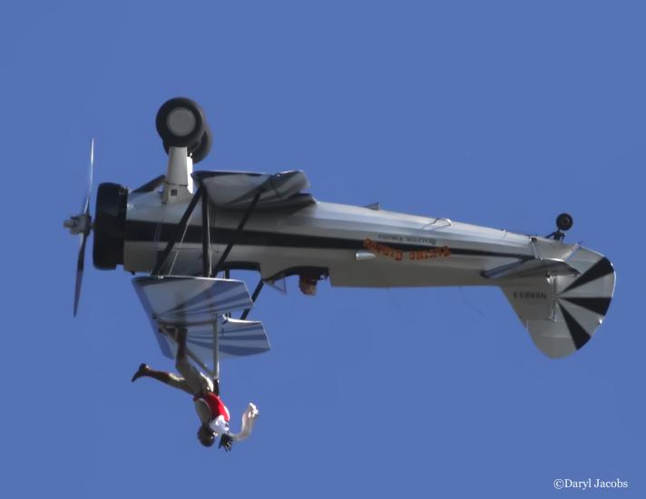 Side view of white and black biplane with single engine flying upside down. A person is depicted as standing on the upper wing of the plane.