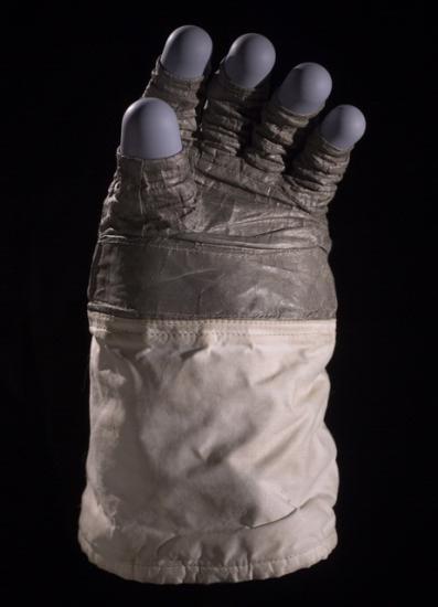 Astronaut glove featuring multiple different fabrics: a leather like fabric for the palm, a purple-colored rubber for the finger tips, and a tan fabric for the sleeve.