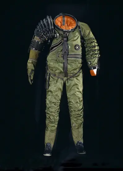 Front view of full-body spacesuit made of dark green-colored material. An opening for the helmet is present and reveals an orange-colored bladder. A black material covers the right shoulder and forearm.