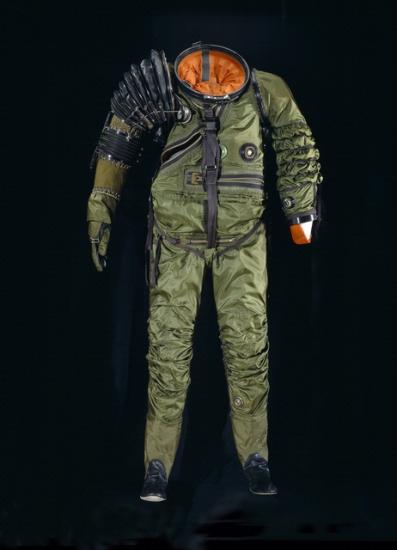 Front view of full-body spacesuit made of dark green-colored material. An opening for the helmet is present and reveals an orange-colored bladder. A black material covers the right shoulder and forearm.