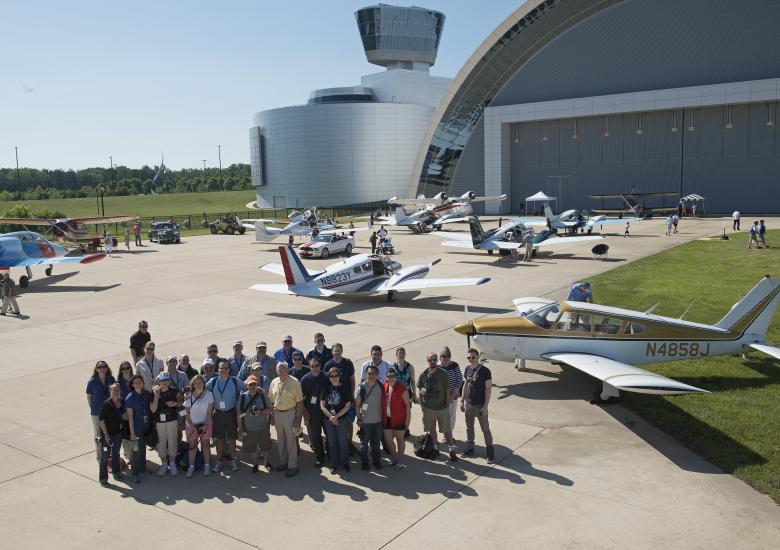 A group of individuals stand on a runway with several planes parked behind them.