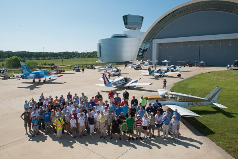 A group of pilots and museum staff pose as a group on a runway with planes behind them.