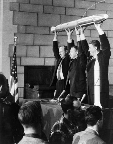 Three people hold a small, pencil-shaped satellite.