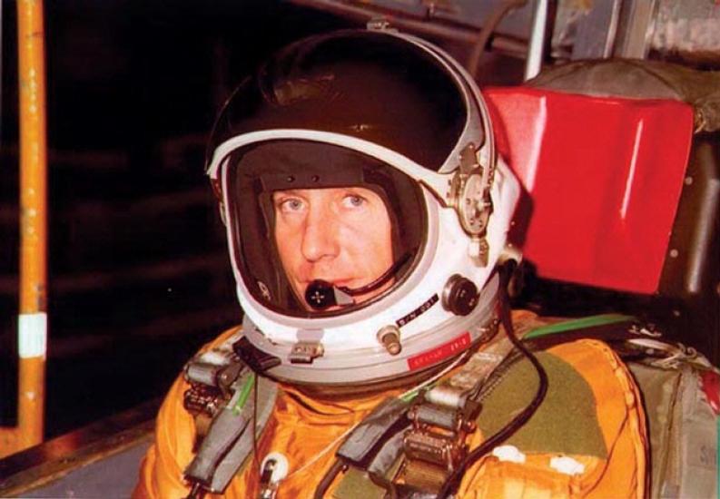 Col. Richard Graham, a white man, sits in an orange and green flight suit and white helmet.