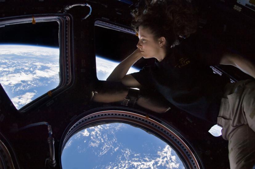 Tracy Caldwell Dyson, a white female astronaut, leans on a window showing the Earth in space.